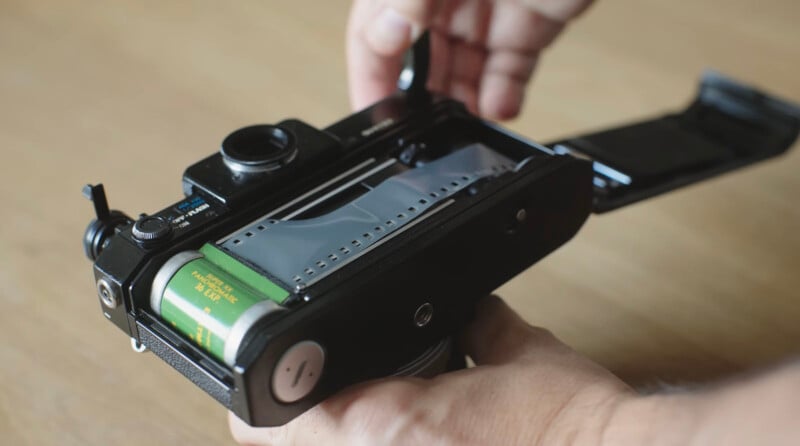 A person’s hands load a roll of 35mm film into the back of a black film camera, with the camera’s door open, showing the film being threaded onto the spool.