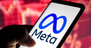 A hand holds a smartphone displaying the Meta logo and name, with a blurred red stock market graph in the background.