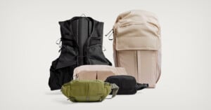 A variety of bags and packs in neutral colors, including a large beige backpack, a black vest-style pack, a green crossbody bag, and two smaller pouches in beige and black, arranged on a plain light background.