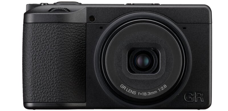 A black compact digital camera with a textured grip on the left, a prominent lens in the center, and minimal buttons or markings on the front.