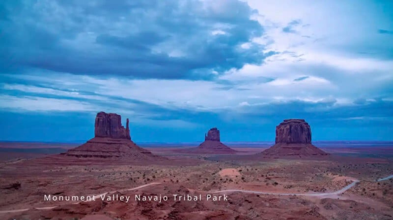 Tres enormes colinas de arenisca se elevan desde el suelo del desierto rojo contra un cielo azul nublado en el Parque Tribal Navajo de Monument Valley. El texto en la parte inferior dice 
