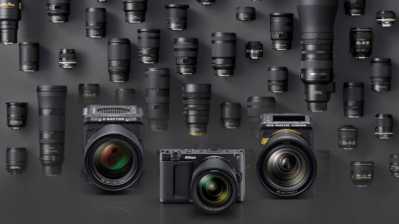 A collection of camera lenses is displayed on a gray surface with three digital cameras in the front row, facing forward among various lens types and sizes arranged in the background.