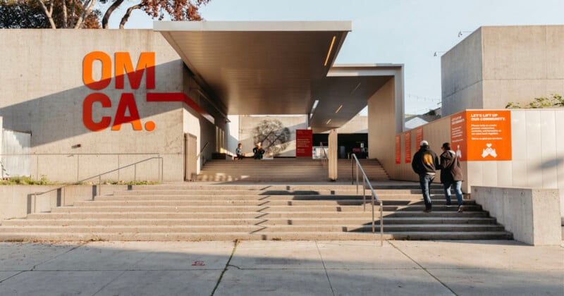 Wide concrete steps lead up to the entrance of a modern museum with large orange "OMCA." letters on the wall and informational signs. A few people walk up the steps under the overhang.