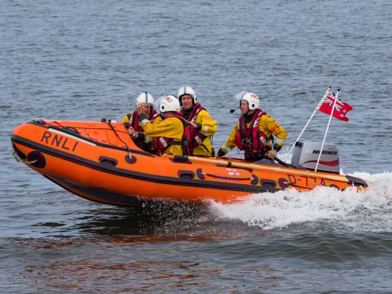 Four people in yellow suits and white helmets ride an orange RNLI rescue boat with a red flag across choppy water, creating a splash.