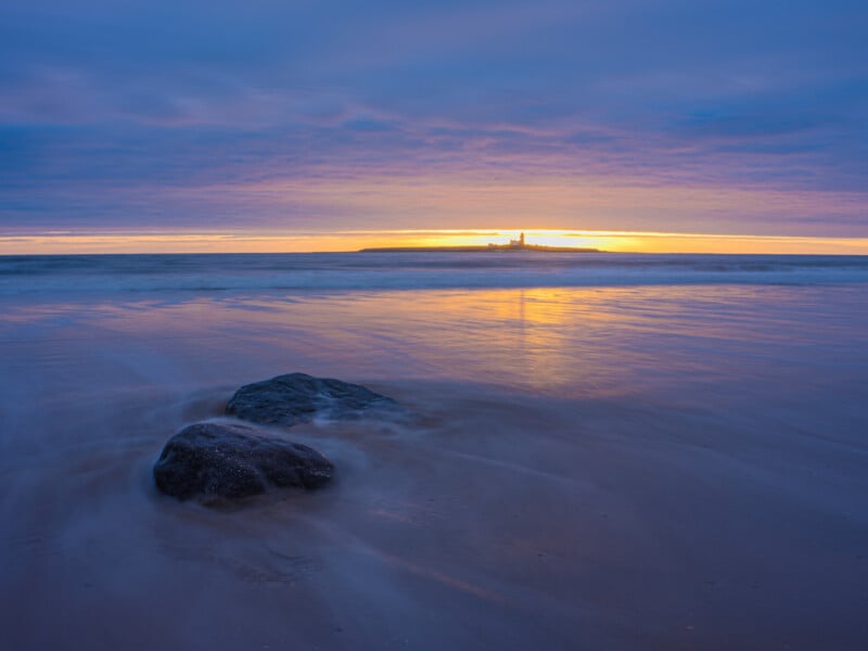 Two rocks on a sandy beach with gentle waves at sunset; the sky is partly cloudy, and distant land with a lighthouse is silhouetted on the horizon under a glowing orange and blue sky.