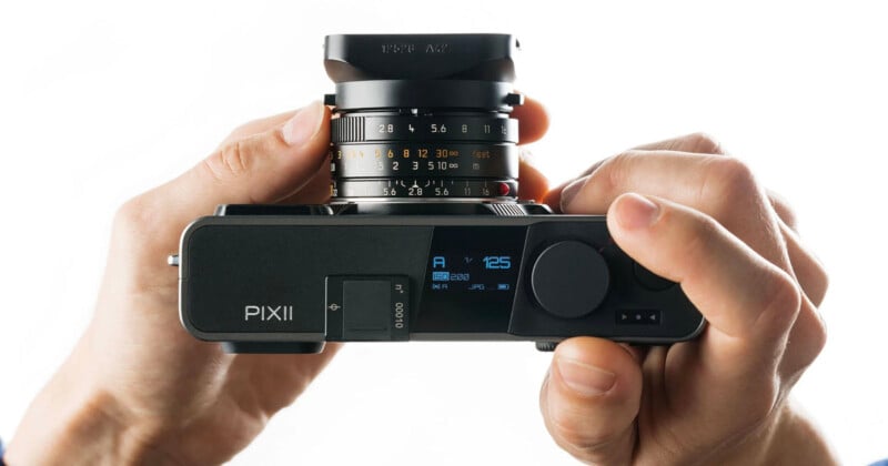 Una persona sostiene una cámara digital PIXII con ambas manos, mostrando una vista superior de la cámara y la lente, con los diales, los botones y la pantalla digital visibles.