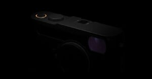 A close-up view of a black camera in low light, highlighting the top dials and a faintly glowing orange ring, with the lens and viewfinder partially visible.