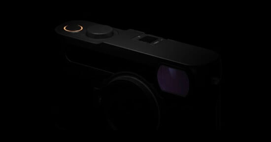 A close-up view of a black camera in low light, highlighting the top dials and a faintly glowing orange ring, with the lens and viewfinder partially visible.