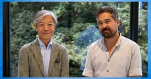 Two men smiling and standing side by side in front of large windows with greenery outside. One wears a gray suit jacket and the other a light-colored casual shirt. The image is framed by a blue border.