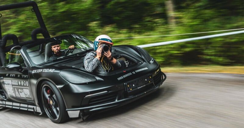 Un hombre con casco toma una fotografía con una cámara mientras está sentado en el asiento del pasajero de un auto deportivo negro que se mueve rápidamente en la carretera, con árboles borrosos en el fondo.