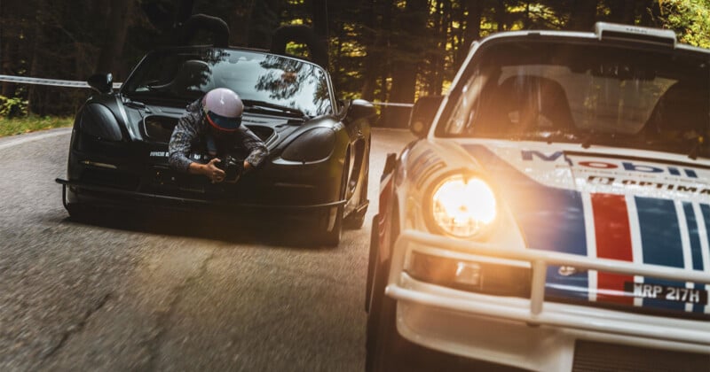 Un hombre con equipo de carreras atado al capó de un auto deportivo negro filmó un auto de rally blanco con franjas azules y rojas, ambos acelerando por una carretera sinuosa en un denso bosque.