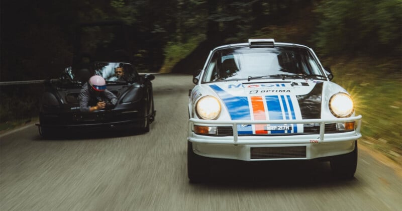 Por una carretera sinuosa circulaba un antiguo Porsche de carreras blanco con franjas azules y rojas, seguido de cerca por un deportivo negro. Un hombre en un coche negro se asoma con una cámara para filmar el Porsche. Paisaje forestal alrededor de la carretera.