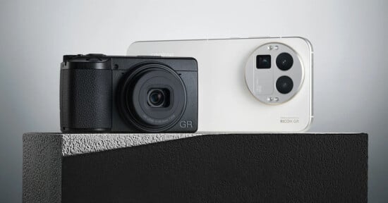 A black compact camera and a white smartphone with triple rear cameras are displayed side by side on a textured gray surface against a minimalist background.
