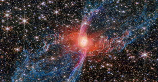 A vibrant nebula with a glowing red center and intricate blue and pink filaments stretches across a star-filled background in outer space. Many bright stars are scattered throughout the scene.