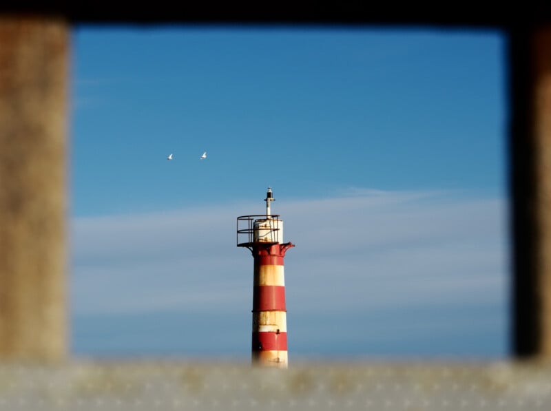 A red and white striped lighthouse stands against a blue sky with two birds flying nearby, framed by a blurred rectangular foreground.