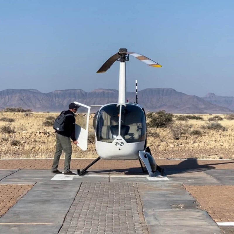A person with a backpack opens the door of a small white helicopter on a paved landing pad in a dry, grassy landscape with distant mountains and a clear blue sky.