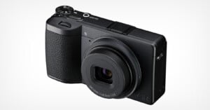 A black compact digital camera with a textured grip, labeled "GR" on the front, featuring a prominent lens and control dials on top, photographed against a plain white background.