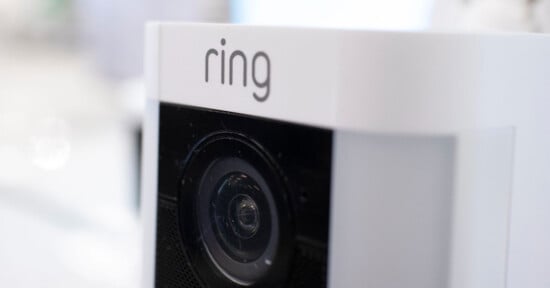 Close-up of a white Ring security camera showing the lens and the "ring" logo at the top. The background is blurred.