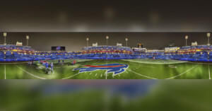 A wide-angle view of an empty football stadium at night, with bright lights and a large buffalo logo at midfield. A few people stand on the turf near the center. Blue seats fill the stands in the background.