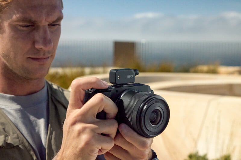A person holds and adjusts a digital camera with a mounted Rode microphone outdoors, with a blurred background of blue sky and wooden structures.