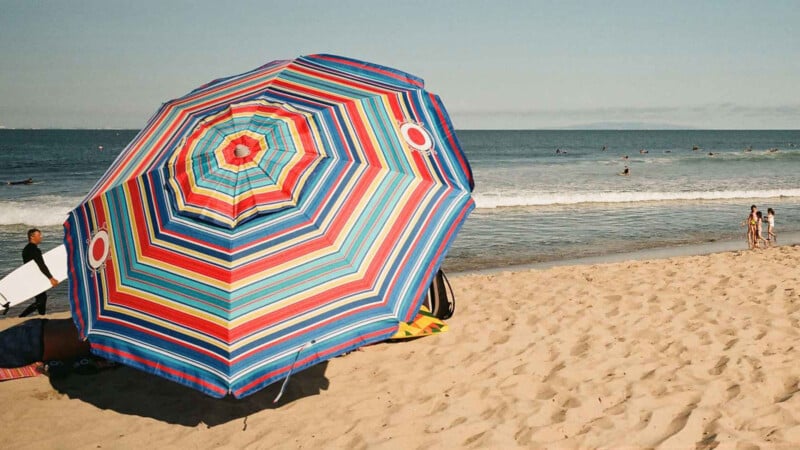 A large, colorful striped beach umbrella shades people relaxing on sandy beach near the ocean. Waves are visible in the background, with a few surfers in the water and people walking along the shore.