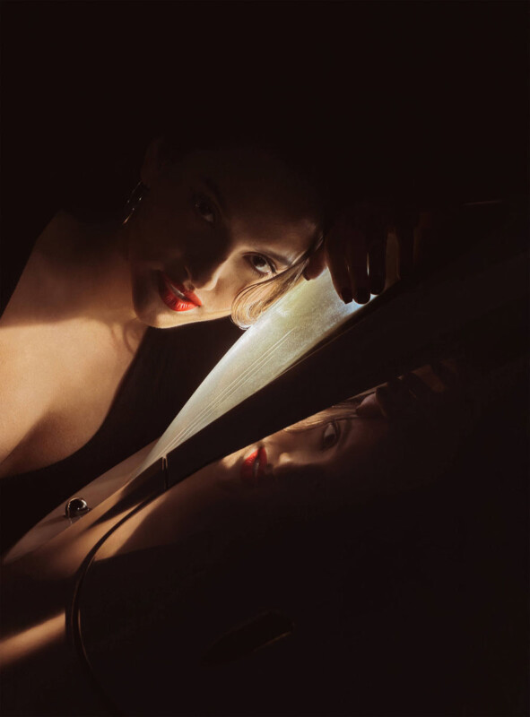 Una mujer que lleva lápiz labial rojo y aretes de aro está sostenida cerca de una superficie reflectante con una iluminación tenue y dramática, proyectando sombras parciales y creando un reflejo cambiante de su rostro debajo.