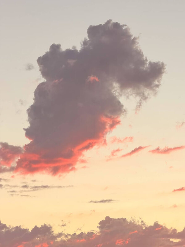 Al atardecer, una nube grande y esponjosa de color gris oscuro y rosa flota en el cielo pastel, con toques de naranja y amarillo cerca del horizonte y nubes más pequeñas dispersas debajo.