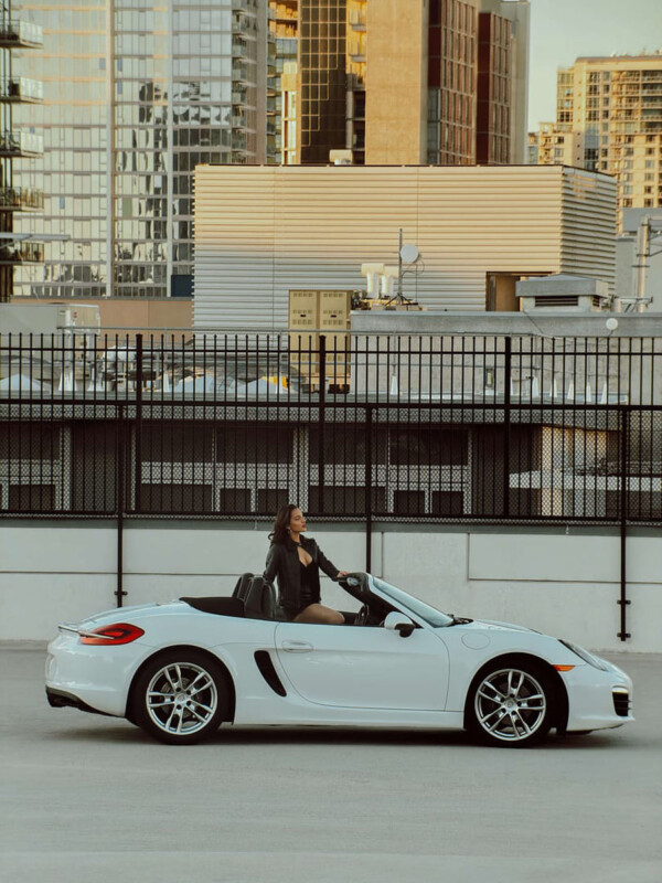 Una mujer sentada en un descapotable blanco con la puerta abierta, estacionado en el techo, con altos y modernos edificios de la ciudad al fondo.