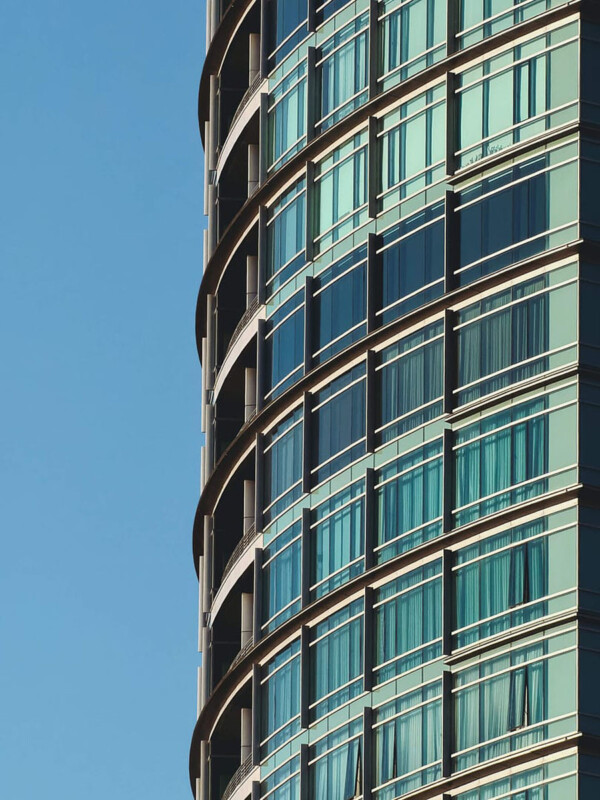 Primer plano de un edificio moderno de gran altura con muchas ventanas de vidrio y balcones que reflejan el cielo azul en su fachada curva. La imagen muestra sólo una parte del edificio, enfatizando sus elegantes líneas arquitectónicas.