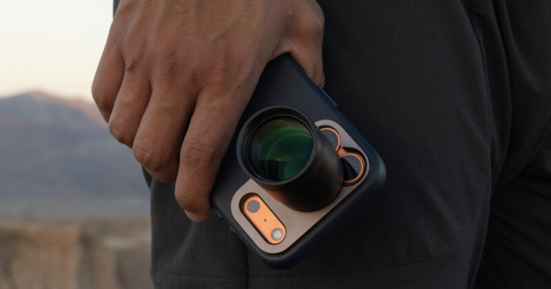 Primer plano de una persona que sostiene un teléfono inteligente con un accesorio de lente de cámara grande, que muestra un paisaje montañoso al aire libre en el fondo.