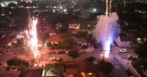 Aerial view of a residential neighborhood at night with fireworks shooting up from the street, illuminating houses and trees while city lights glow in the background.