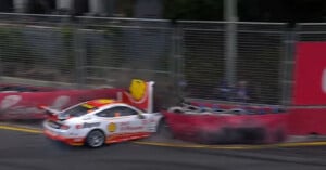 A white and orange race car crashes into a barrier on a race track, sending up smoke and debris near a fence and tire wall.