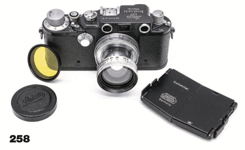 A vintage Leica camera with a silver lens, a yellow filter, a lens cap, and a black folding viewfinder, all arranged on a white surface. The number 258 appears in the lower left corner.