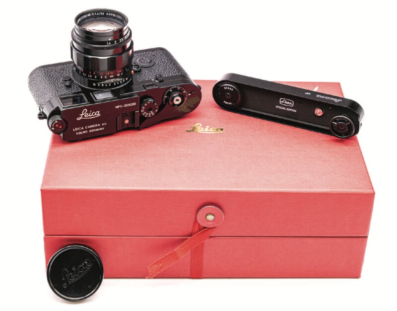 A black Leica camera with lens, a separate light meter, and a lens cap are displayed on a closed red Leica box with a buttoned strap. The Leica logo is visible on the box and the camera.