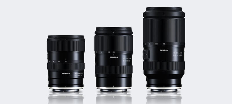 Three black Tamron camera lenses of varying heights are standing upright side by side on a reflective surface, with visible focal length markings and model details on each lens. The background is light gray.