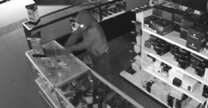 A person wearing a hooded jacket and gloves is smashing a glass display case inside a store, surrounded by shelves of electronics. The scene is captured in black and white, likely from a security camera at night.