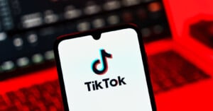 A smartphone displays the TikTok logo on its screen, with a blurred laptop in the background. The scene is illuminated by a red light.