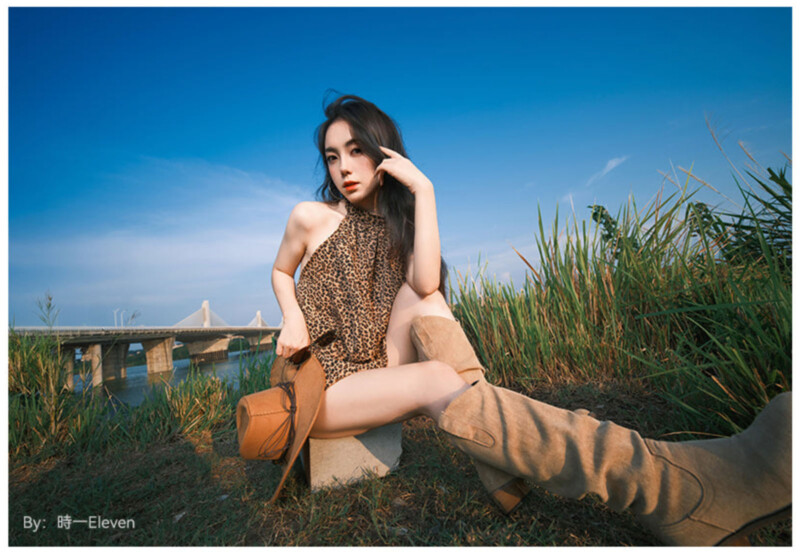 Una mujer con un vestido con estampado de leopardo y botas color canela posa al aire libre en el césped cerca de altos juncos, con un río y un puente bajo un cielo azul claro al fondo.