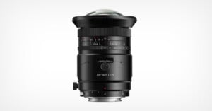 A black tilt-shift camera lens is displayed upright against a plain white background, showing focus and aperture markings, adjustment knobs, and a wide, domed front glass element.