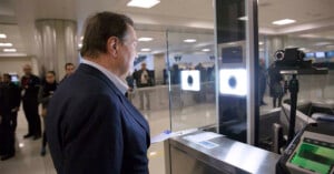 A man stands in front of a biometric scanner at an airport security checkpoint. Several people and staff are visible in the background of the brightly lit terminal.