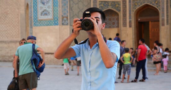 A young man in a light blue shirt takes a photo with a camera in front of an ornate building, while several people walk and stand in the background.