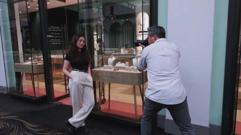 Un fotógrafo capturó una fotografía de una mujer posando frente al escaparate de una joyería. La mujer, vestida con un top negro y pantalón blanco, tenía las manos en los bolsillos mientras el fotógrafo se agachaba y le apuntaba con la cámara.