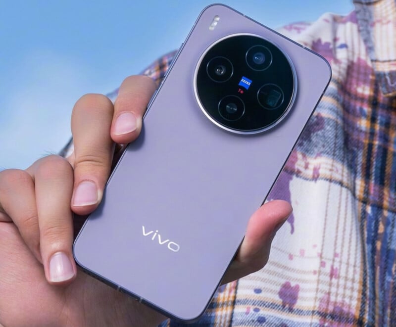 Una persona sostiene un teléfono inteligente Vivo de color violeta con un gran módulo de cámara circular y cuatro lentes en la parte posterior. El hombre lleva una camisa a cuadros con un cielo azul de fondo.