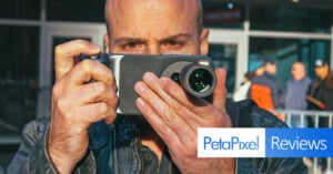 A man holds a smartphone with an attached camera lens, aiming it toward the viewer. The background is slightly blurred, showing people outdoors. The image features "PetaPixel Reviews" text in the bottom right corner.