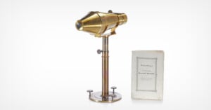 A vintage brass microscope on a metal stand beside an old booklet with a gray border and German text on the cover, both placed against a white background.
