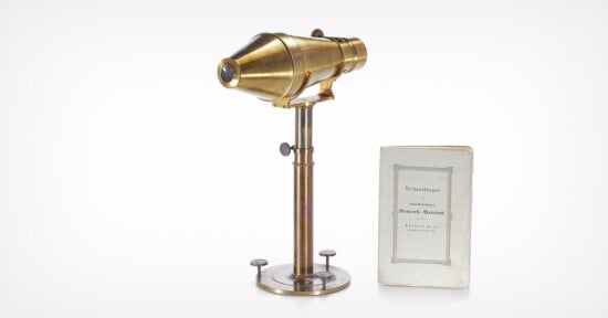 A vintage brass microscope on a metal stand beside an old booklet with a gray border and German text on the cover, both placed against a white background.