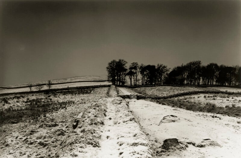 A snow-covered rural landscape with a path leading to a wooden gate, flanked by stone walls and leafless trees in the distance beneath a cloudy sky.