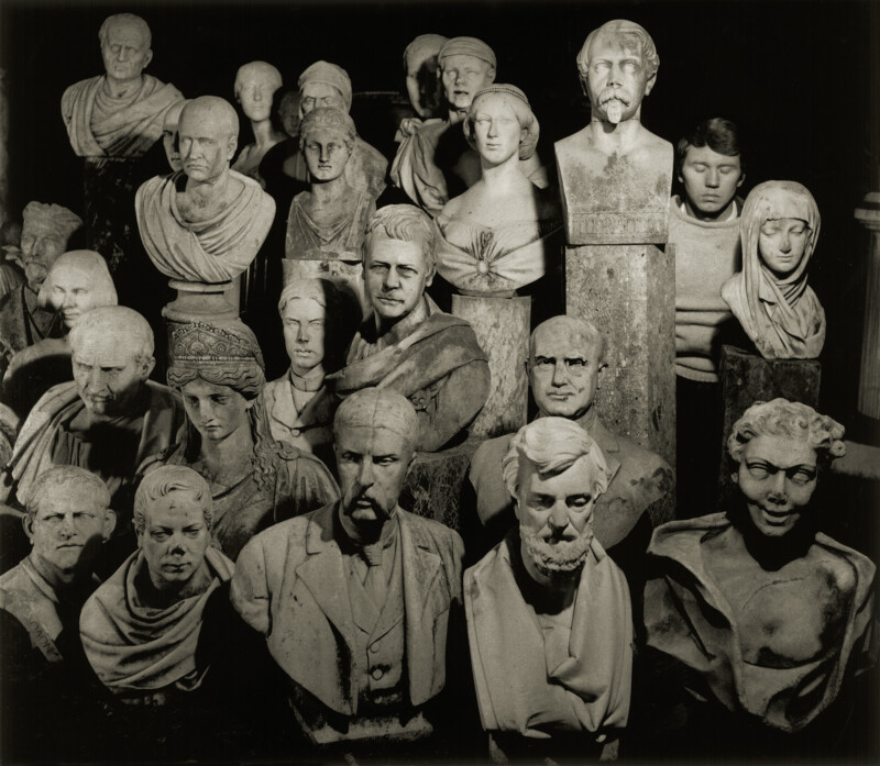 A group of various marble and plaster busts arranged closely together, depicting men and women with different facial expressions, hairstyles, and clothing styles, all lit dramatically against a dark background.