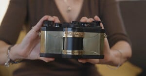 A person in a brown top holds a vintage-style camera with gold and black detailing, displaying it toward the viewer. The camera has a unique, boxy design and the person's hands frame it on each side.