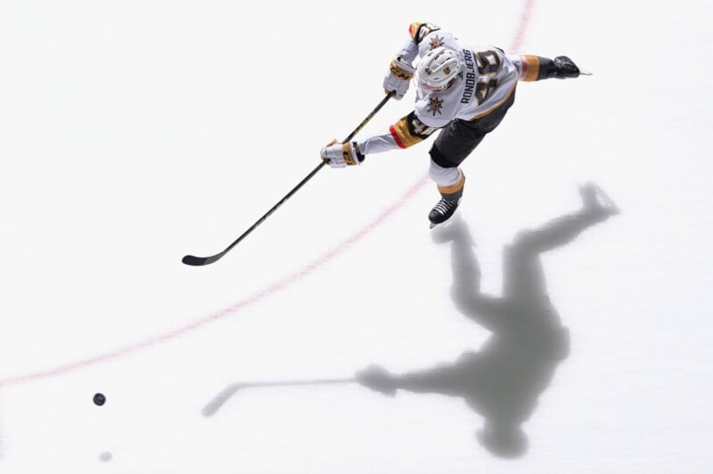 A hockey player in a white and gold jersey takes a shot on the ice, with his stick extended and the puck in front of him. His shadow is clearly visible on the ice below.